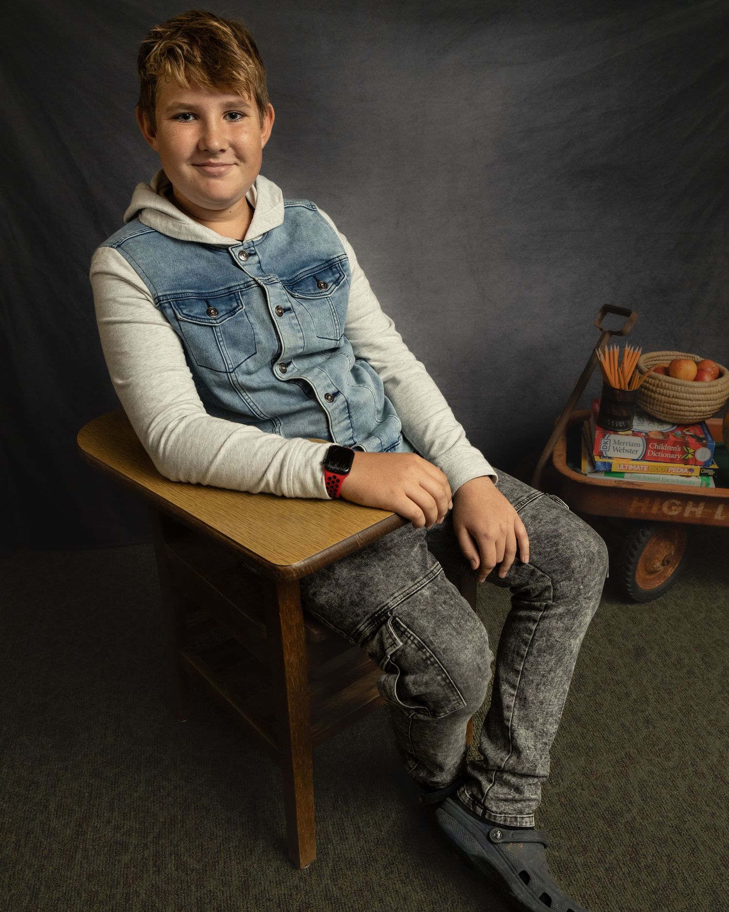 Student seated at a vintage school desk wearing a denim vest and gray jeans against a dark backdrop.