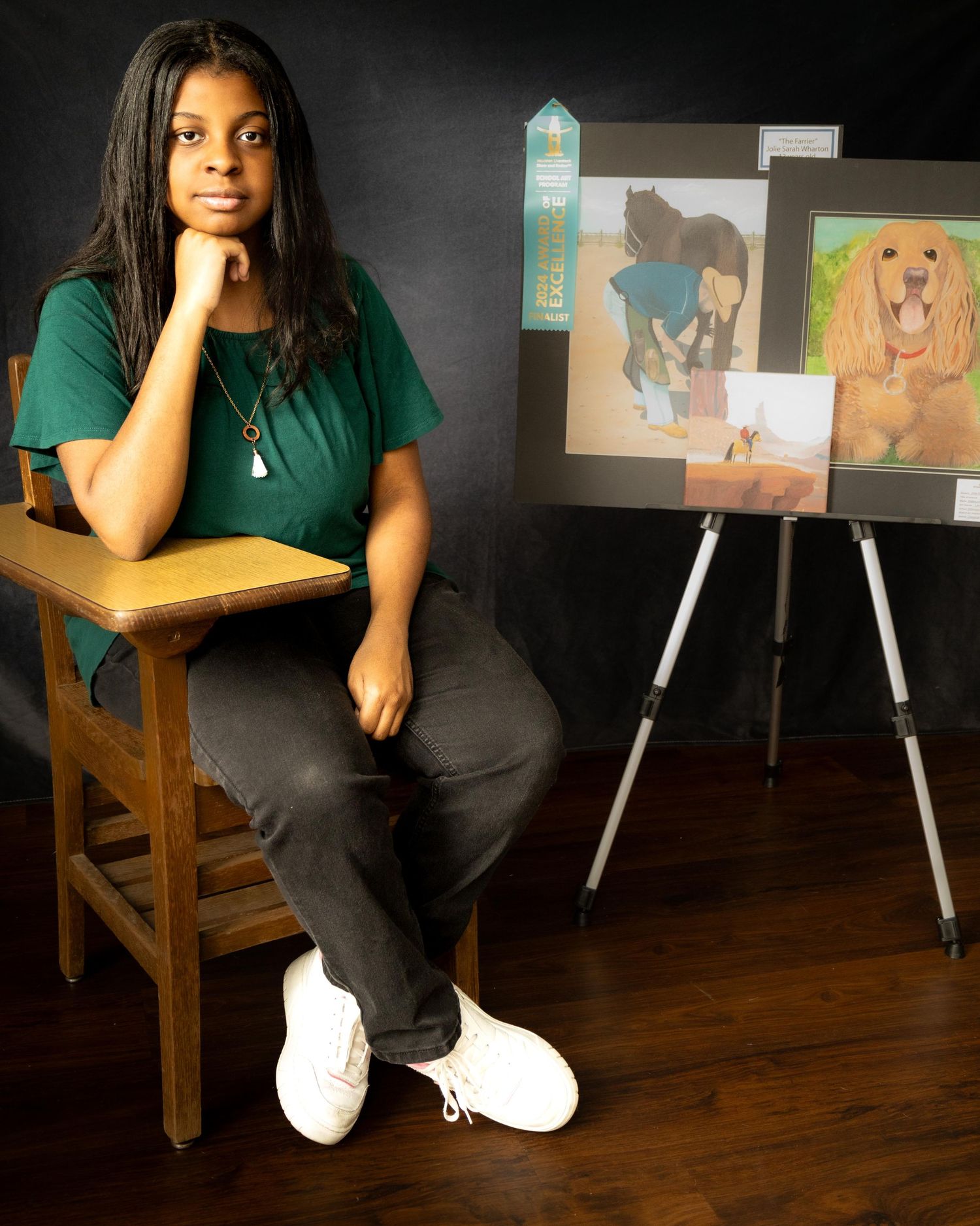 Artist sits thoughtfully on wooden stool next to easel displaying colorful pet portraits in studio setting.