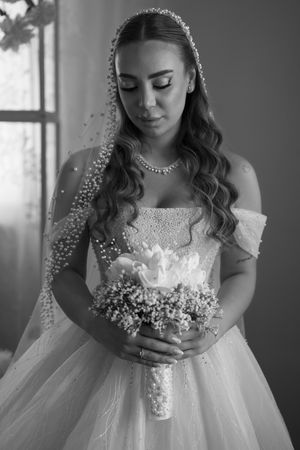 A bride in an elegant white gown with long curls holds a bouquet while looking down in a black and white portrait.