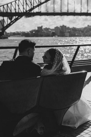 Couple sitting on bench overlooking Sydney Harbour Bridge in romantic wedding photo.