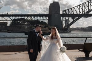 Wedding couple poses under iconic Sydney Harbour Bridge with dramatic cloudy sky.