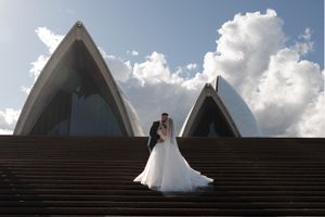 Wedding photo session on steps of Sydney Opera House with fluffy clouds overhead.