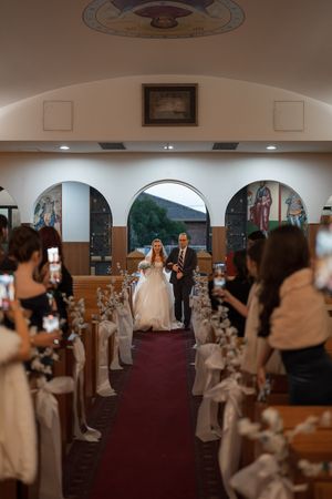 Wedding ceremony taking place in an ornate church with arched doorways and guests capturing the moment on phones.