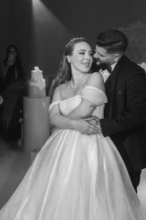 Black and white portrait of couple sharing intimate dance with wedding cake visible in background.