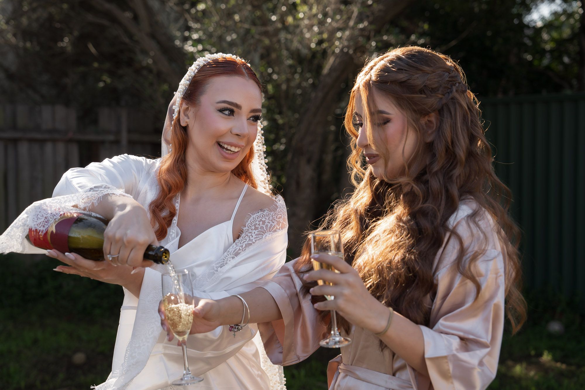 Two women in matching white robes share champagne and laughter during pre-wedding celebrations.