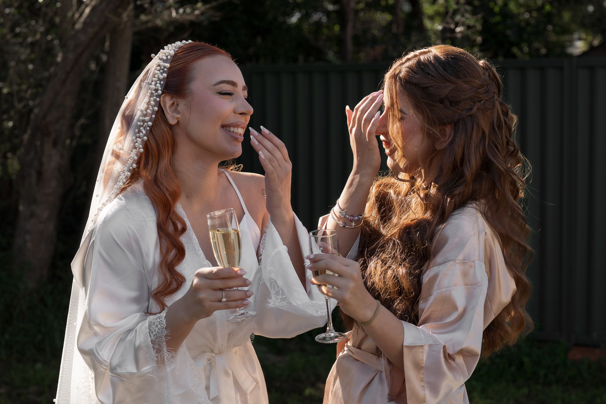 Two friends in white robes share joyful moments while holding champagne glasses outdoors.