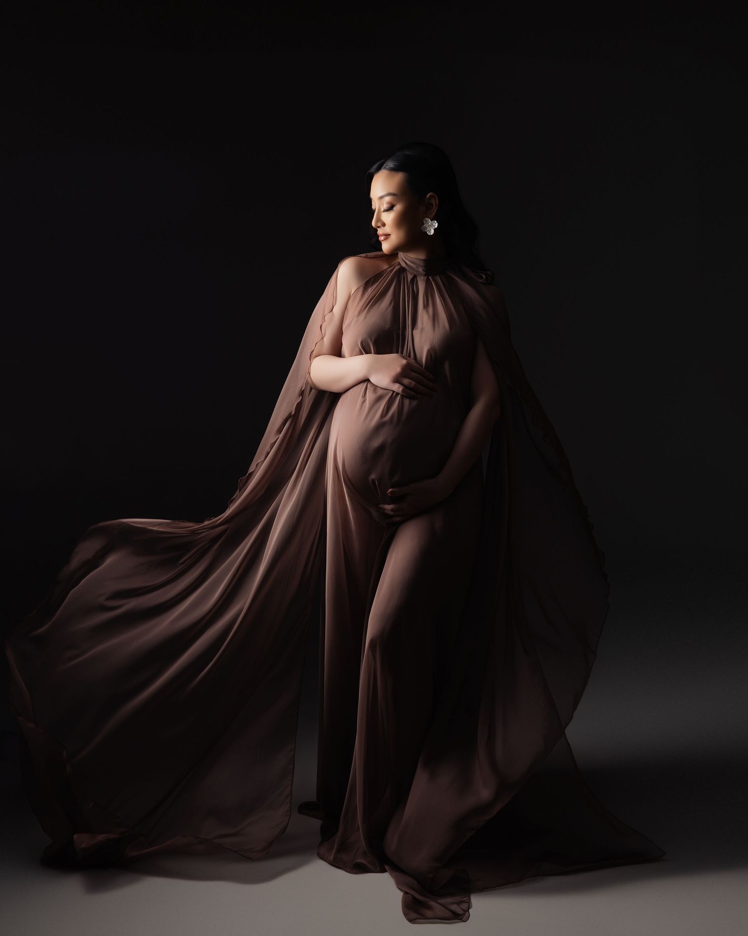Dramatic silhouette photography of a woman in flowing brown fabric dress posing elegantly against dark background.