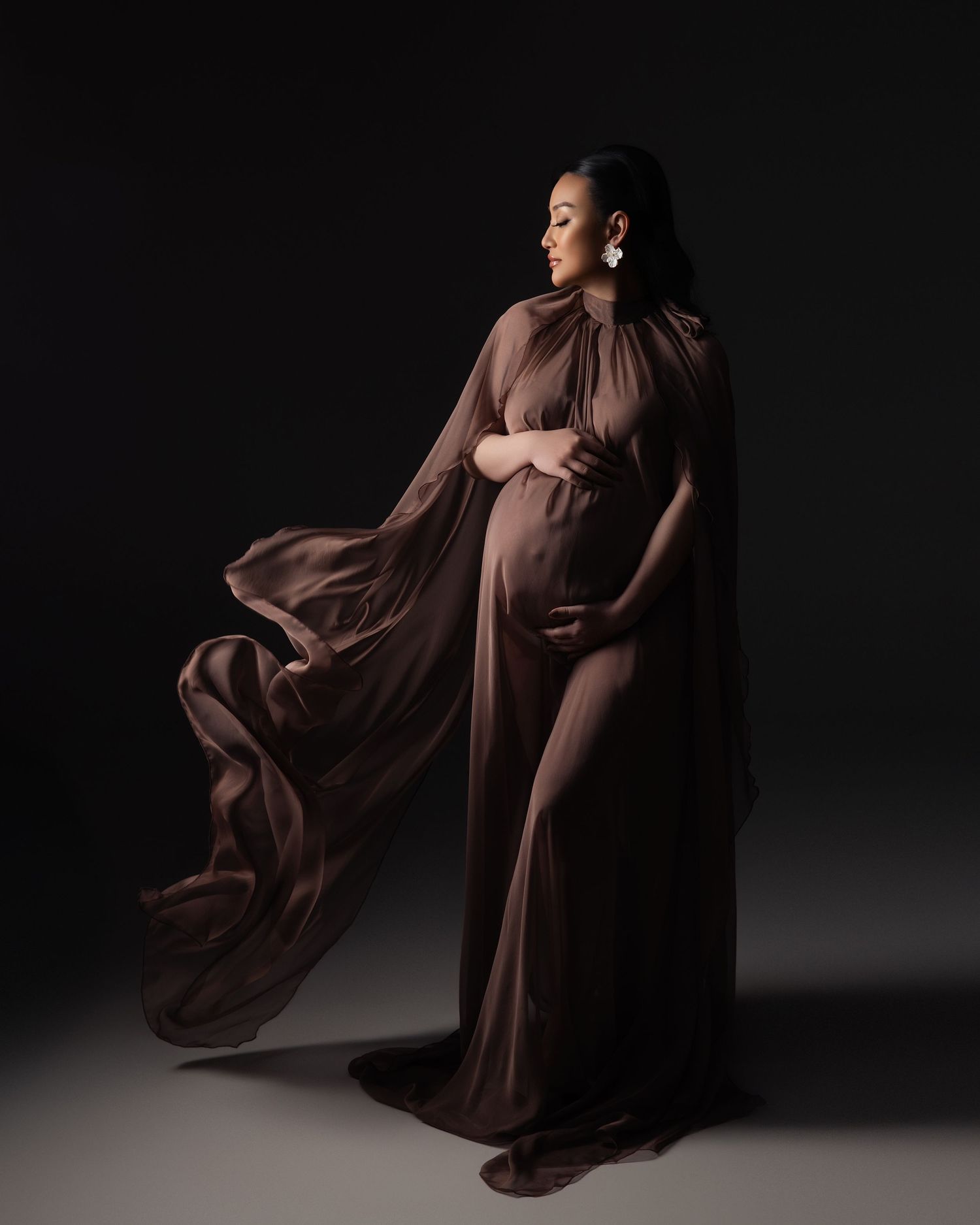 An elegant maternity portrait showing a flowing brown dress against a dark backdrop with dramatic lighting.