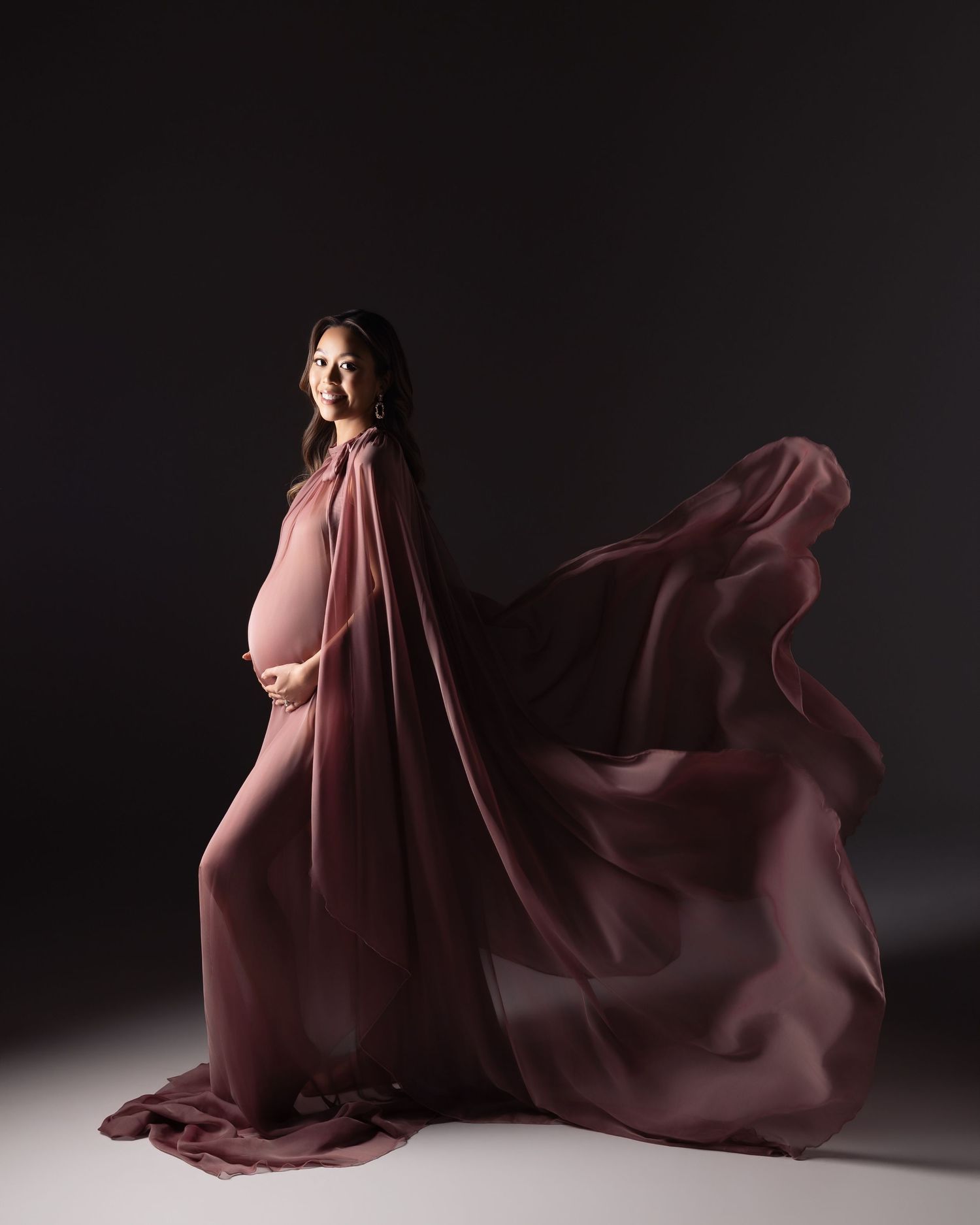 Pregnant woman in flowing mauve chiffon gown with dramatic cape, posed against dark studio background.