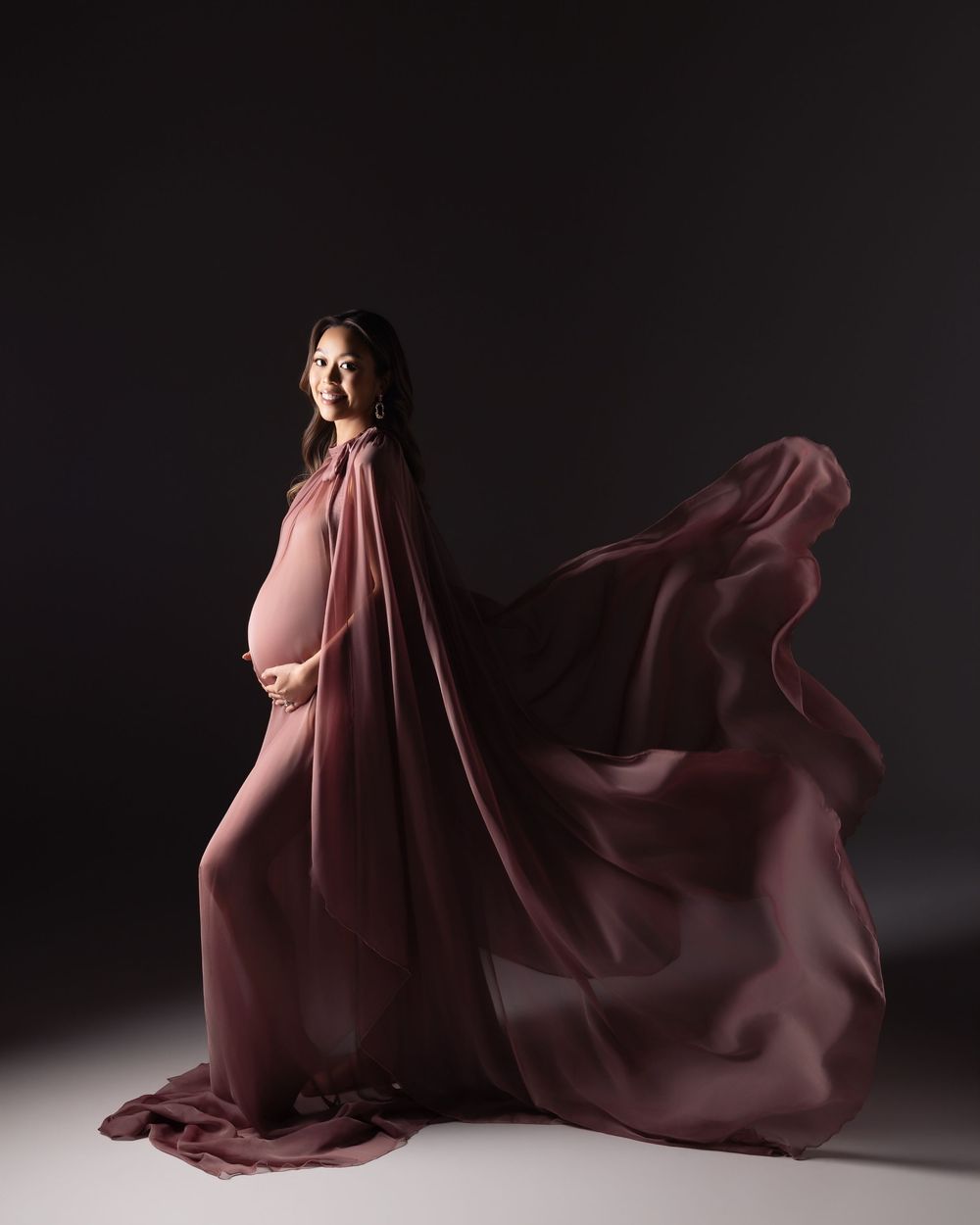 Pregnant woman in flowing mauve chiffon gown with dramatic cape, posed against dark studio background.