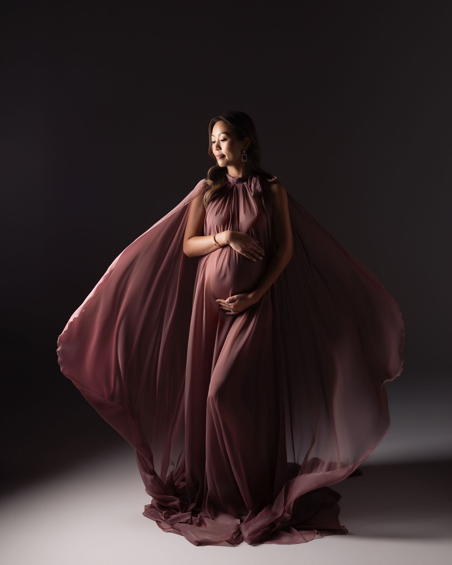Pregnant woman in flowing mauve chiffon cape gown poses gracefully against dark studio background.