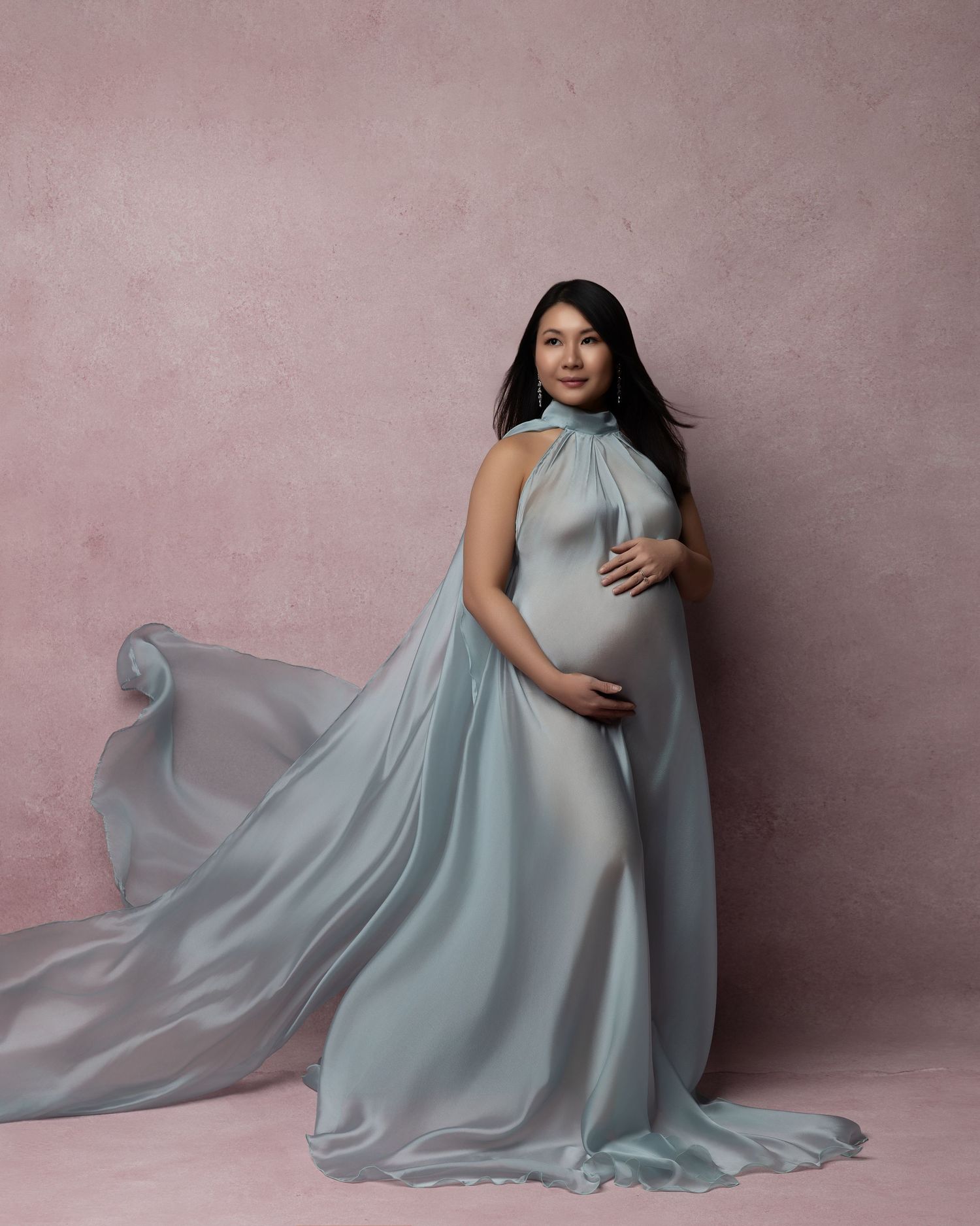 A flowing light blue maternity gown with elegant draping fabric captures movement in a studio setting with pink backdrop.