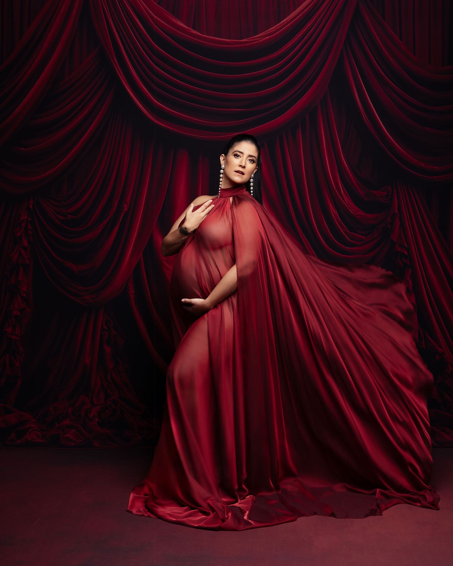 A figure in a flowing red gown poses dramatically against dark red velvet curtains in a theatrical setting.