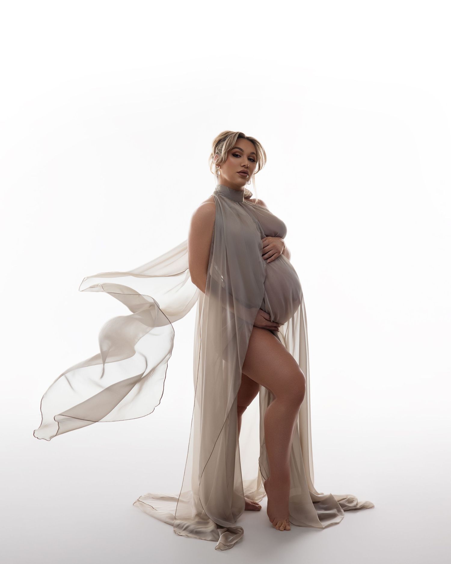 Elegant maternity photo featuring flowing sheer fabric draping around a pregnant figure against a bright white background.