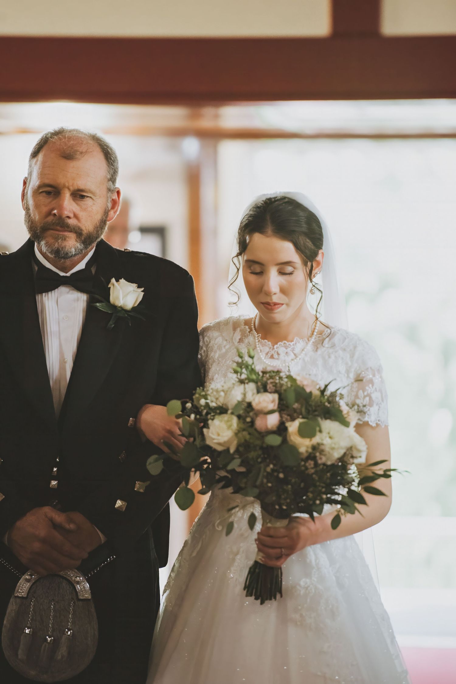 A traditional Scottish wedding moment with the father of the bride in a kilt and bride holding a bouquet of roses and greenery.