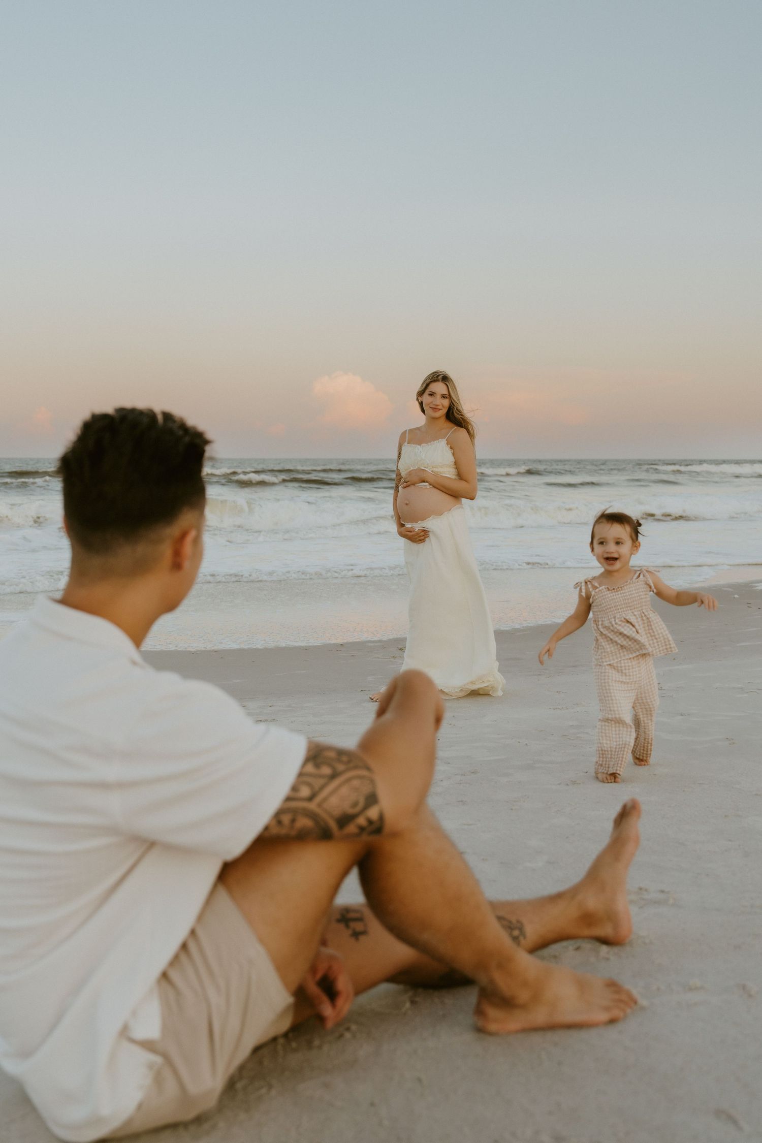 A couple in light-colored attire strolls along a sandy beach path with palm trees and golden sunlight in the background.