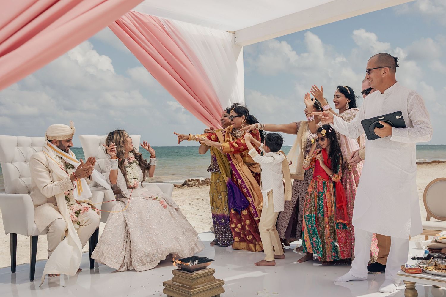 Group celebrates at a beach wedding ceremony with colorful traditional attire under pink draped decor and blue ocean views.