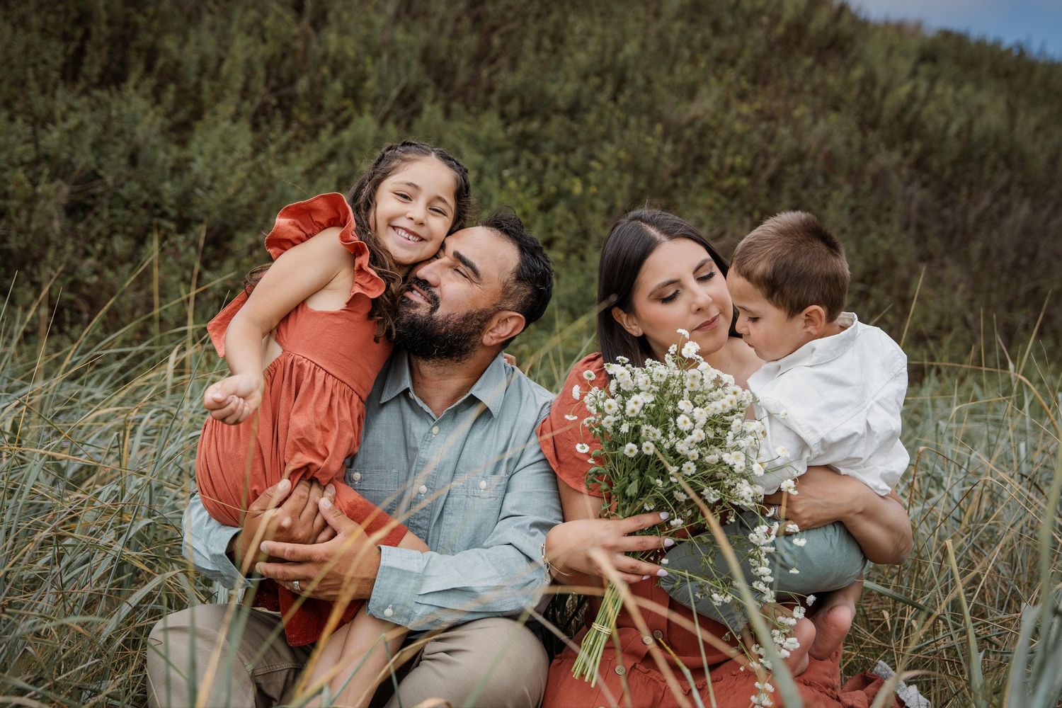 Family wearing orange and blue sits together in a sun-drenched meadow during an outdoor portrait session.