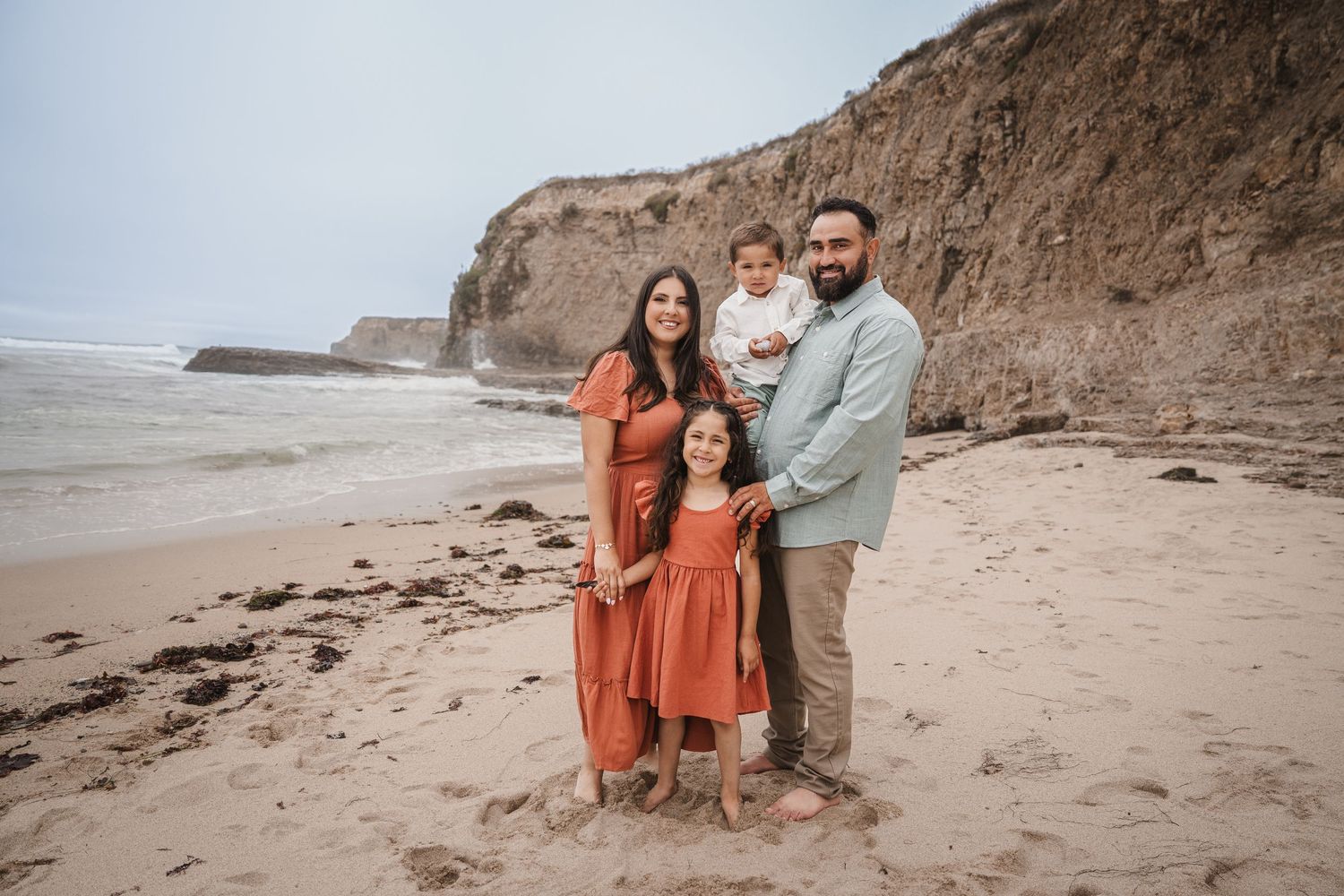 A group wearing coordinated rust-colored outfits poses together on a sandy beach with dramatic coastal cliffs in background.
