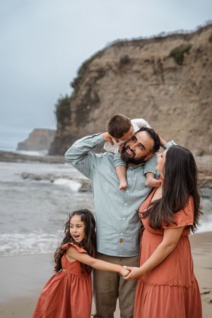 Family in coordinated outfits enjoying a candid moment together on a beach with dramatic coastal cliffs in background.