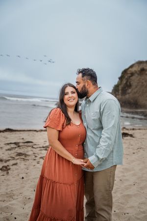 A couple stands lovingly on a cloudy beach with cliffs in the background and birds flying overhead.