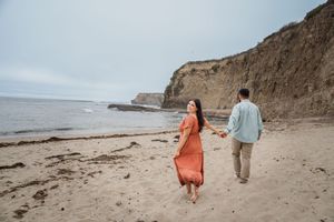 Two people walk hand in hand along a scenic beach with cliffs in the background during golden hour.