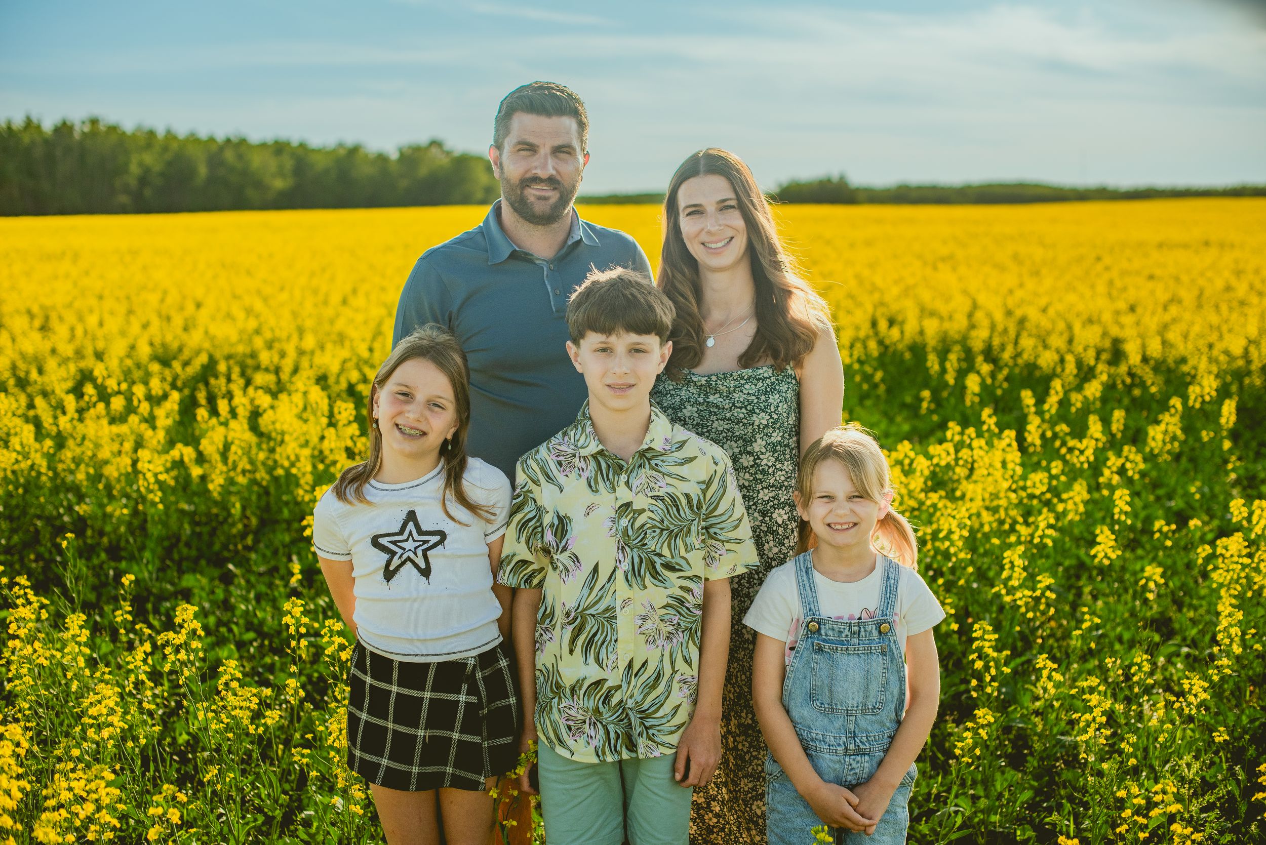 Family poses together in front of vibrant yellow canola field on a sunny summer day.