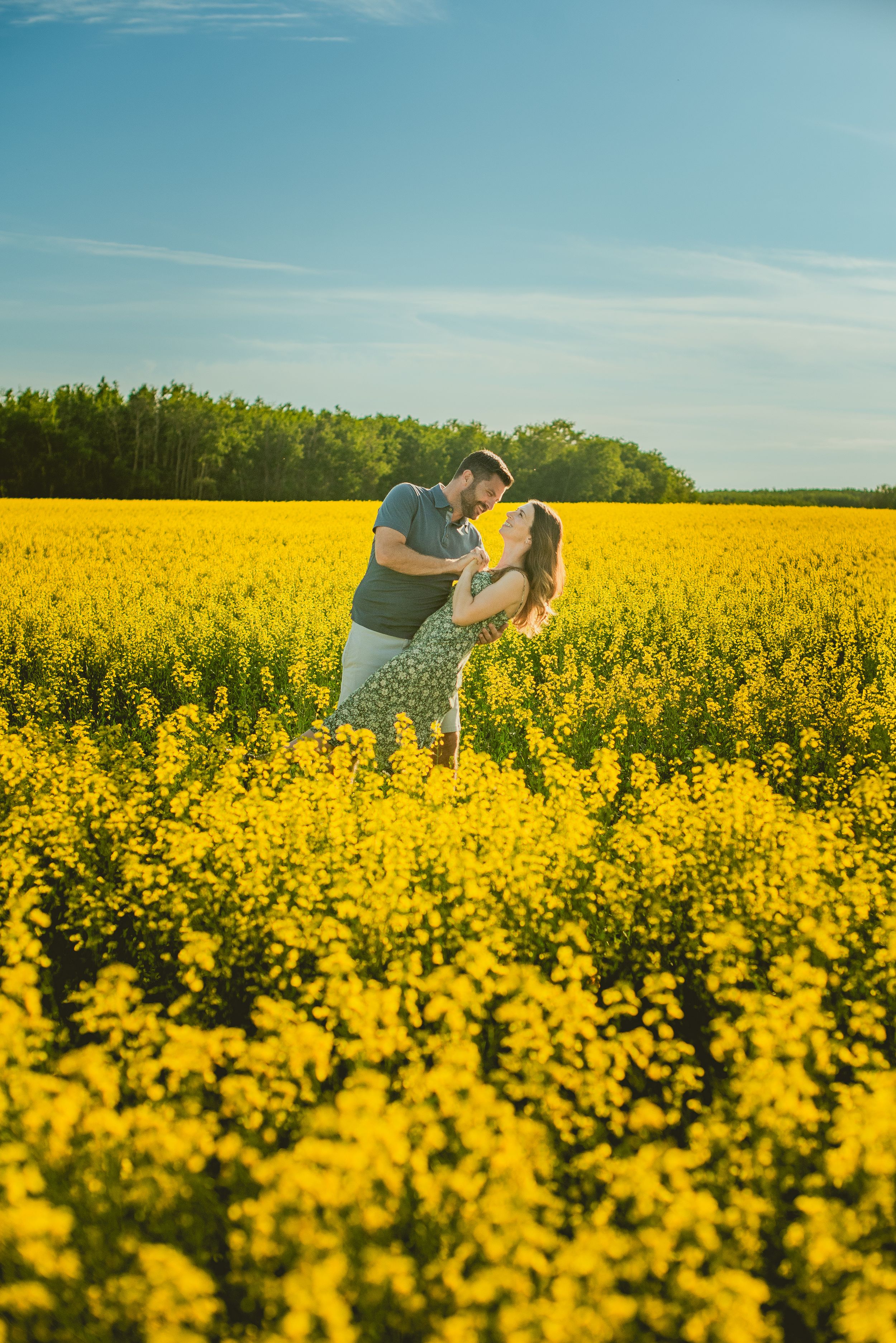A couple embraces in a vibrant yellow canola field on a sunny day with forest and blue sky in the background.