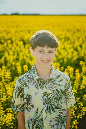 A child wearing a tropical print shirt poses in a vibrant yellow canola field on a sunny day.