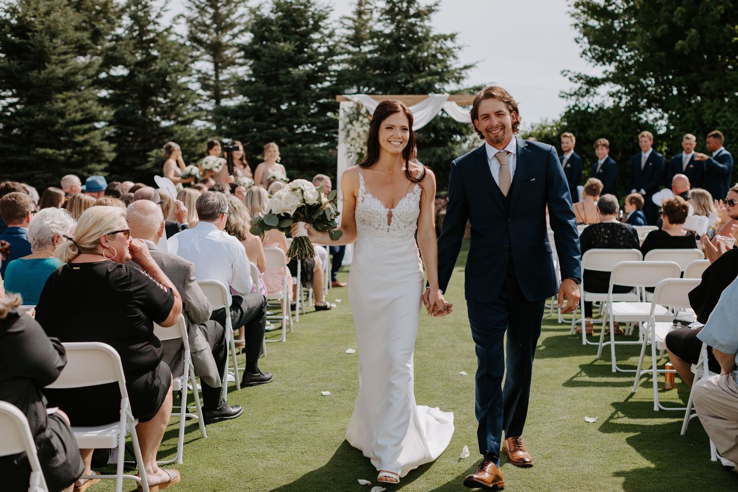 Newlyweds walk down the aisle while wedding guests smile from white chairs on either side.