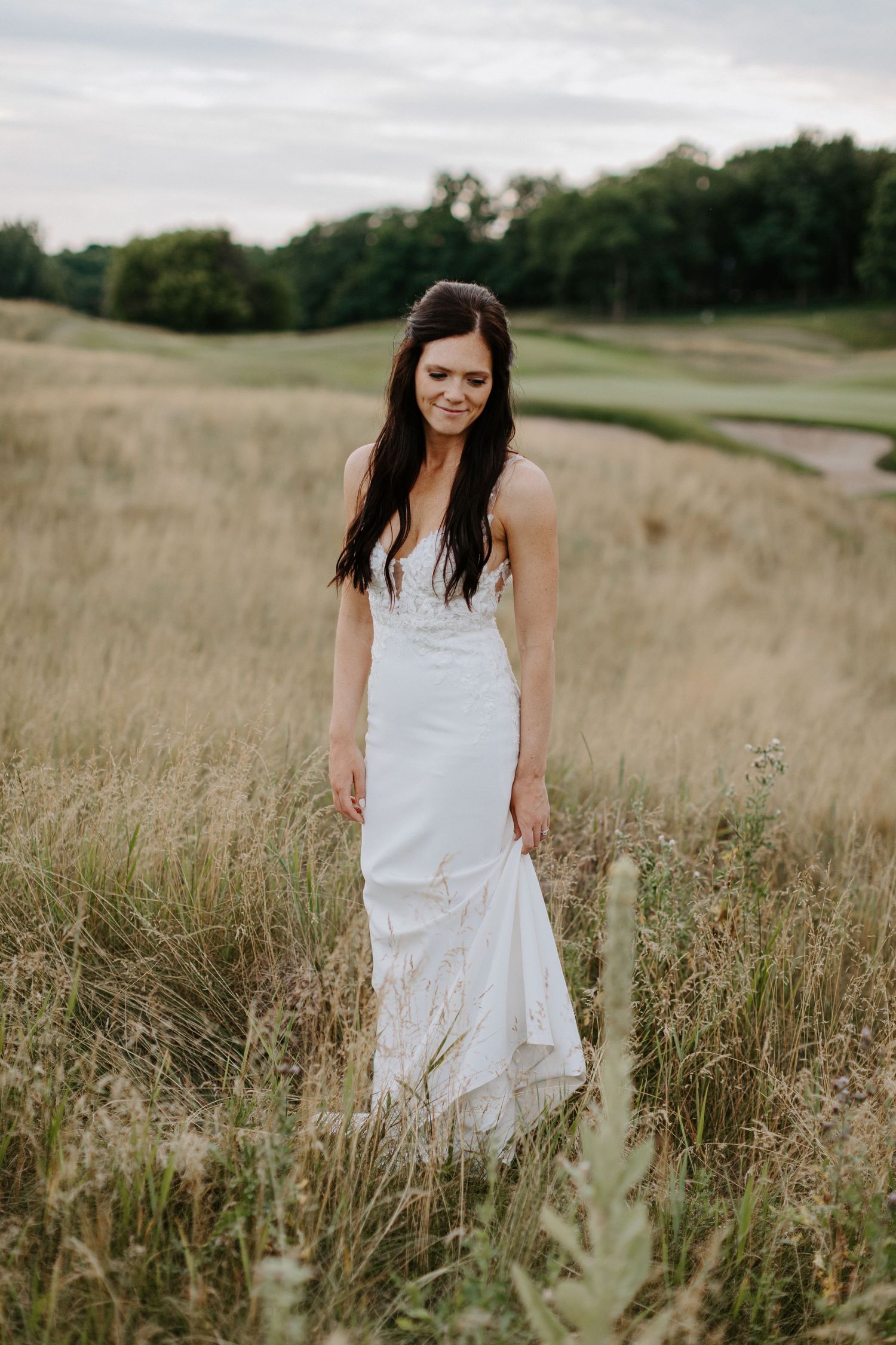 Elegant bride in fitted white dress standing alone in golden wild grass field.
