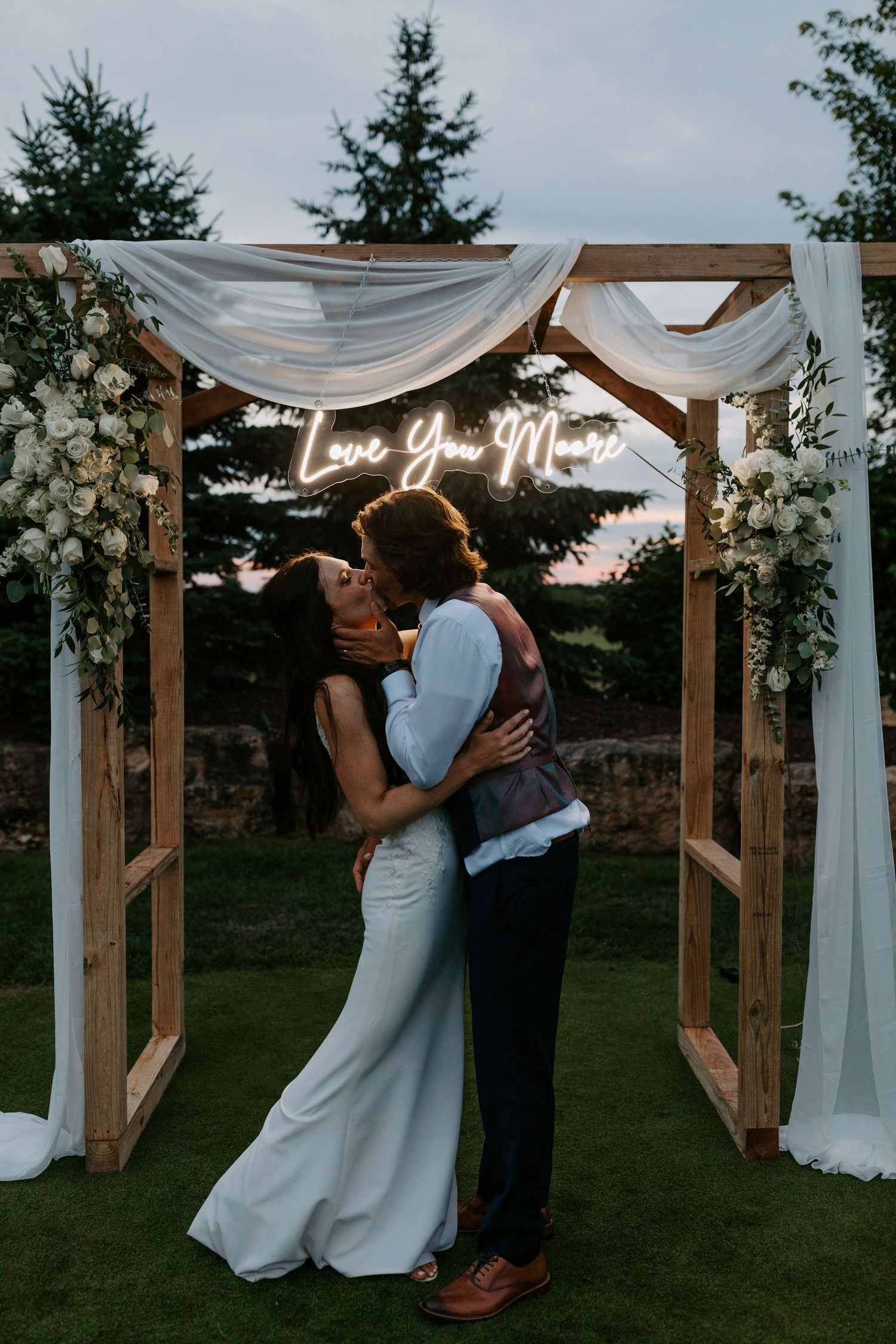 Romantic wedding couple embraces under wooden arch with neon 'Love You More' sign and white fabric draping.