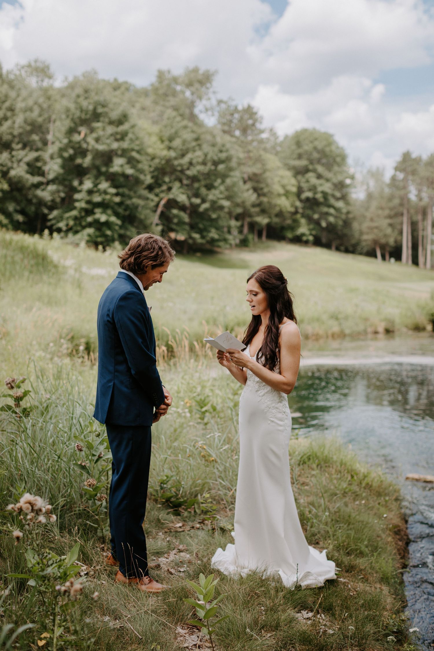 Couple in wedding attire exchanging vows by a serene pond surrounded by tall grass.