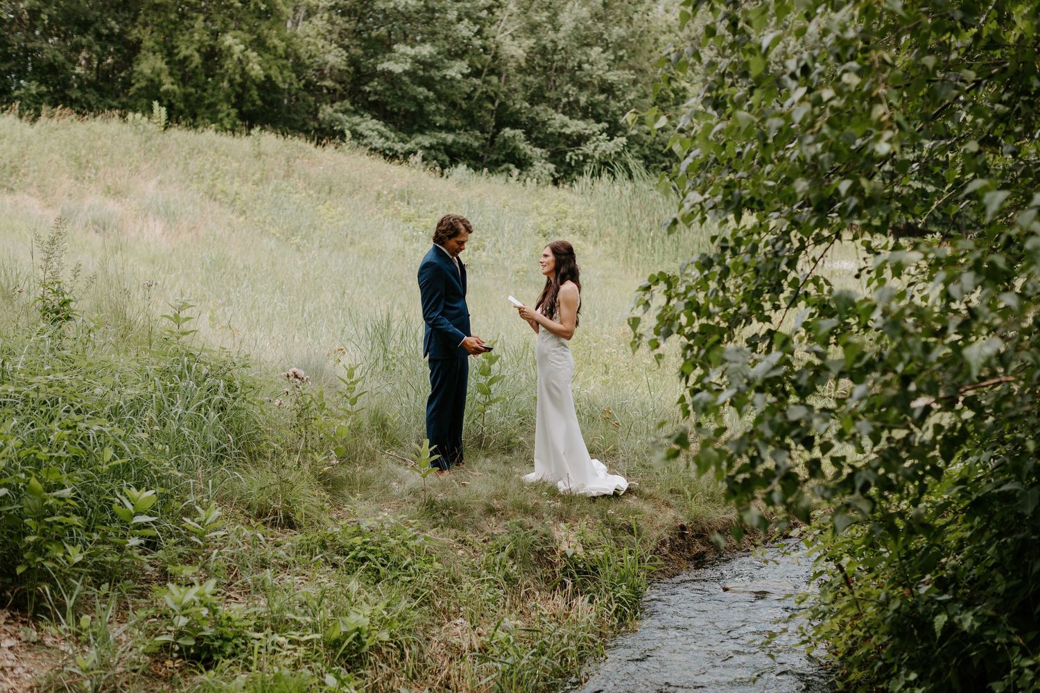 Wedding couple stands intimately by a stream in a natural meadow setting.