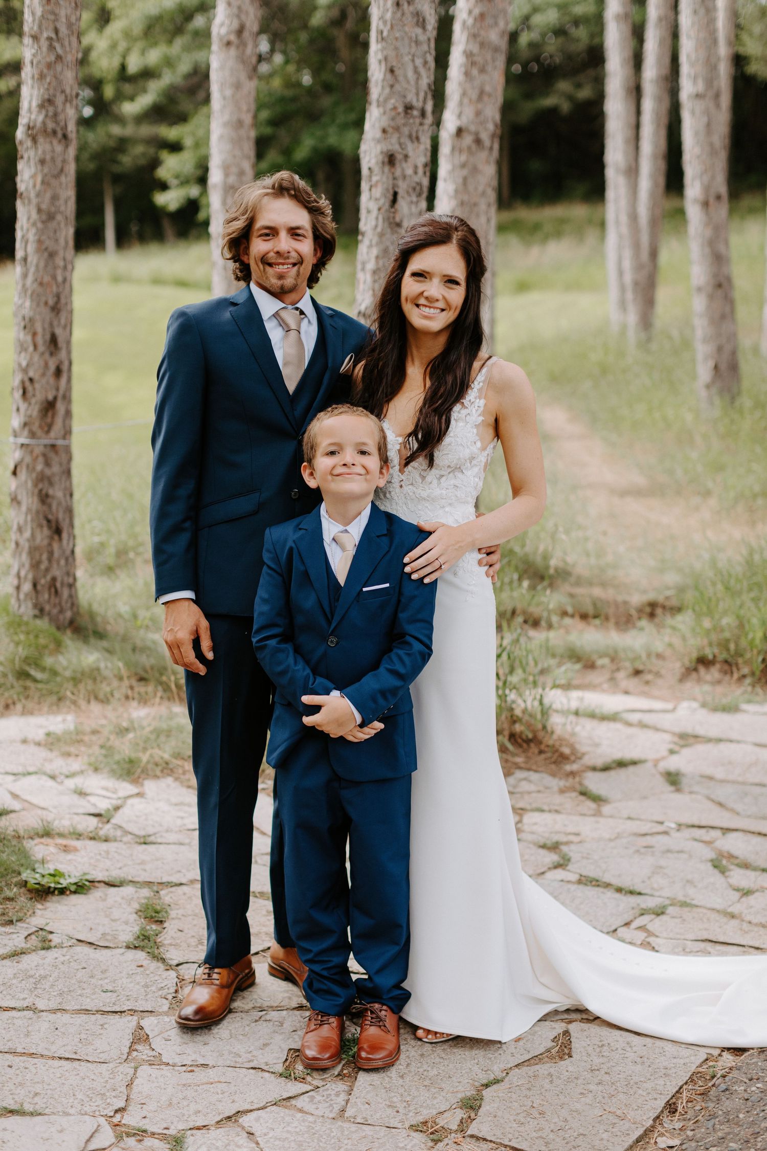 Wedding portrait with couple and young boy in matching navy suits against pine tree backdrop.