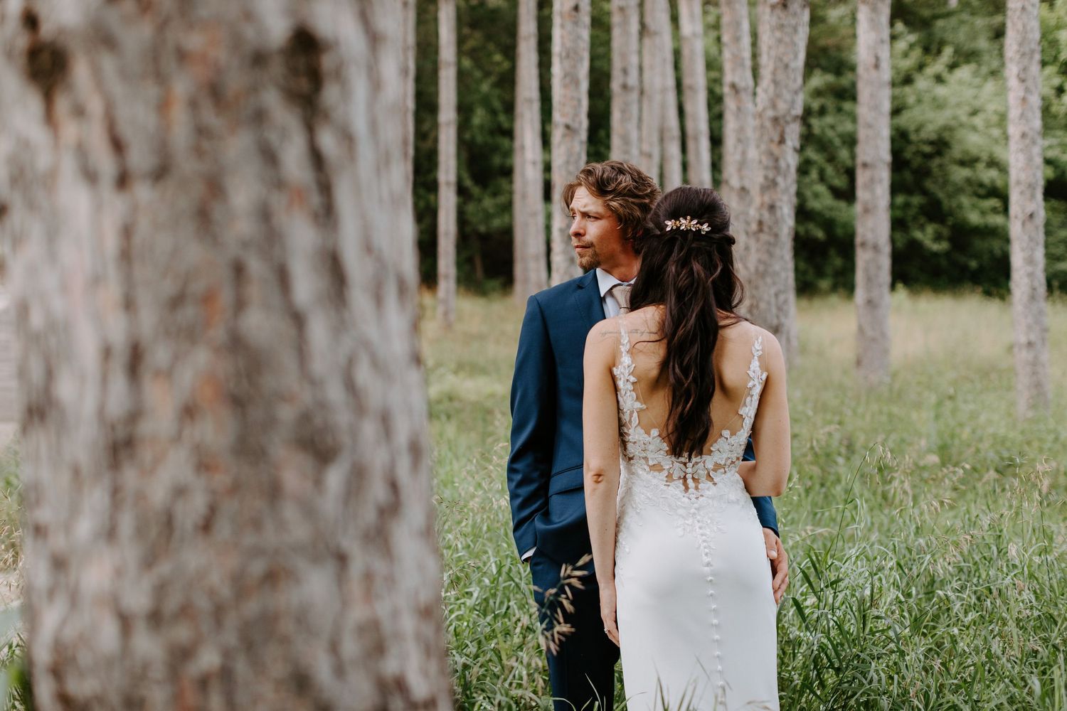 Back view of couple in wedding attire standing among tall pine trees in natural setting.