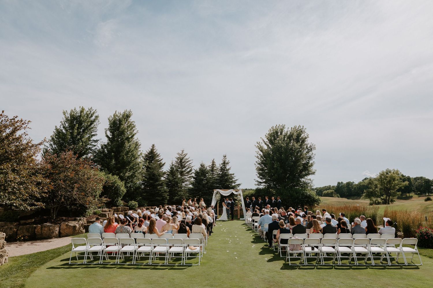 Outdoor wedding ceremony setup with rows of white chairs on a grassy lawn surrounded by evergreen trees.
