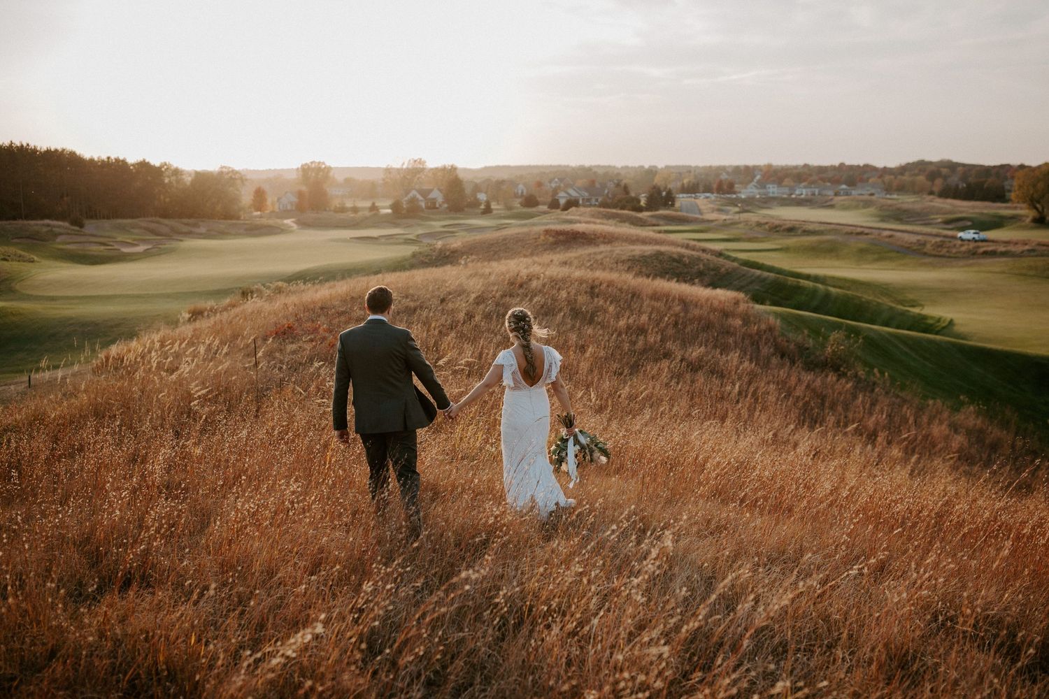 Couple walking through golden grass on hilltop overlooking golf course at sunset.