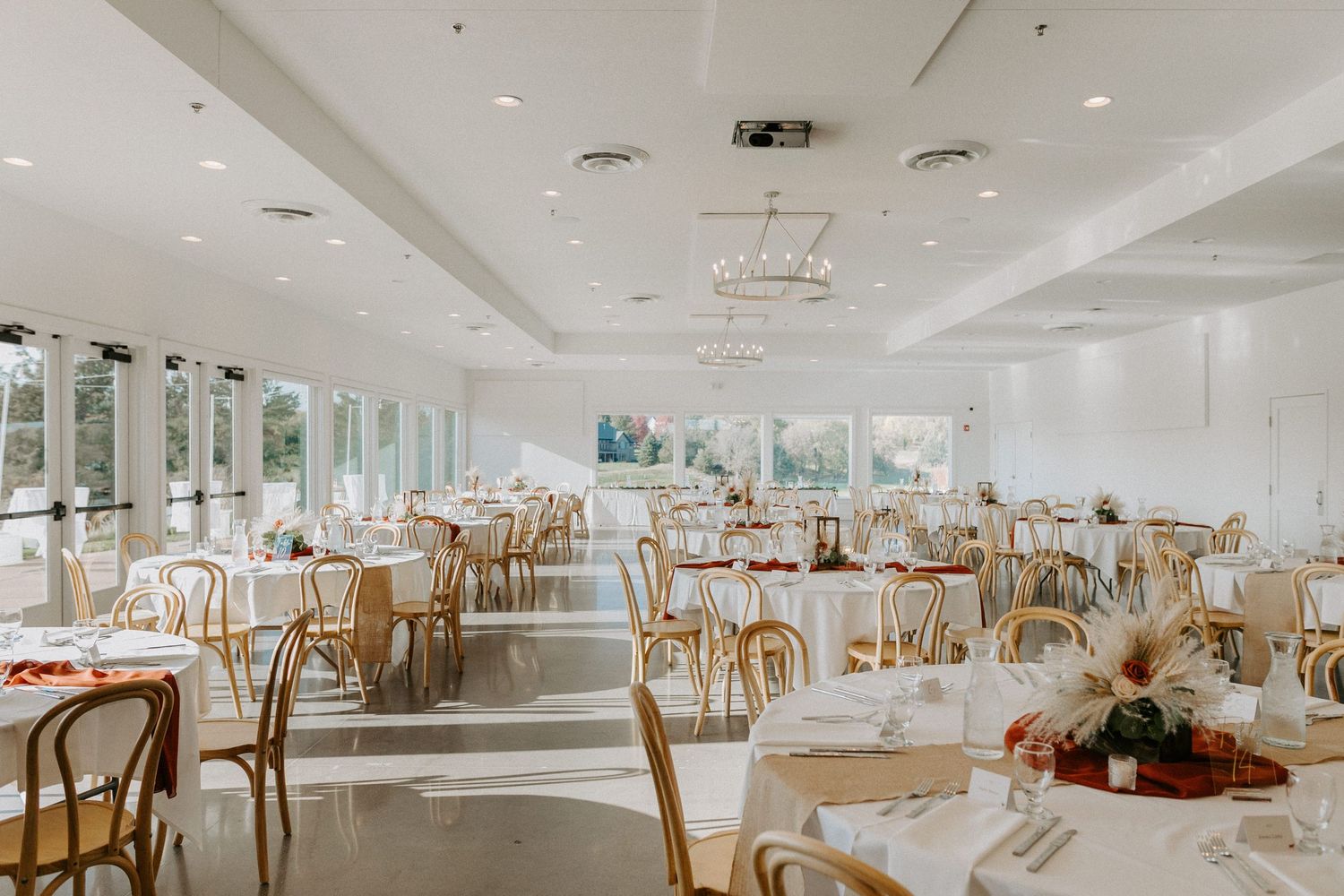 Spacious white wedding reception hall decorated with round tables and wooden chairs.