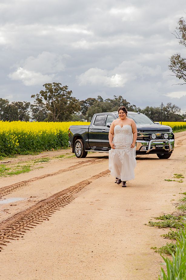 Car tracks lead through a dirt path beside a bright yellow canola field on a cloudy day in rural Australia.