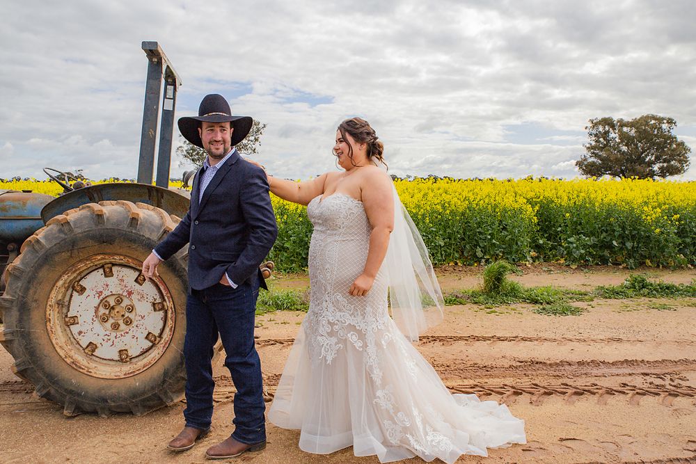A couple poses in wedding attire next to a large tractor with bright yellow canola fields in the background.