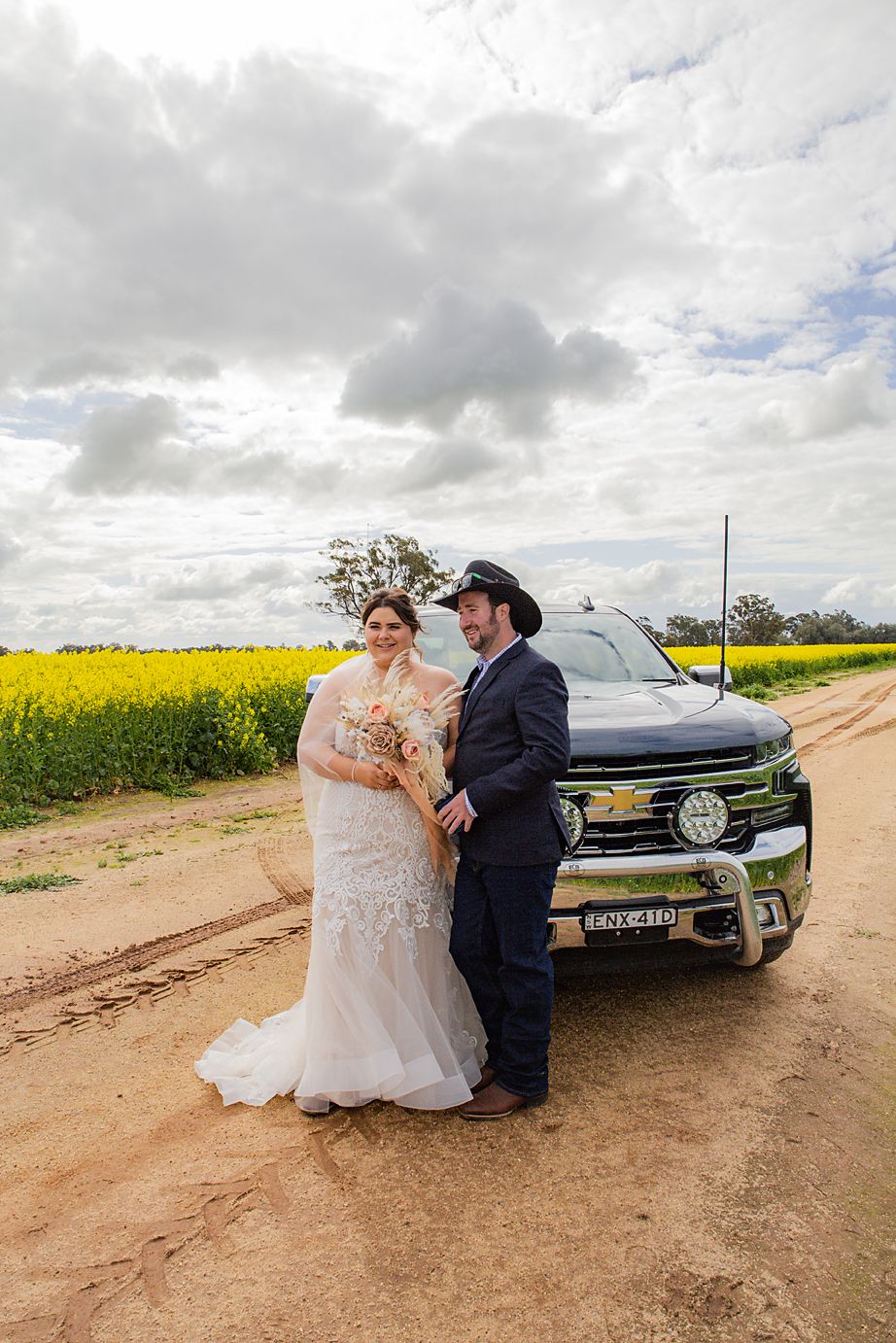 Wedding couple poses together on a dirt road beside vintage car with blooming yellow canola fields in background.