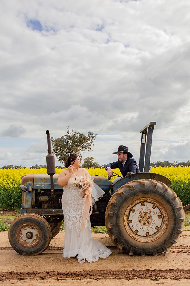 Couple poses with vintage blue tractor in field of bright yellow canola flowers under cloudy sky.
