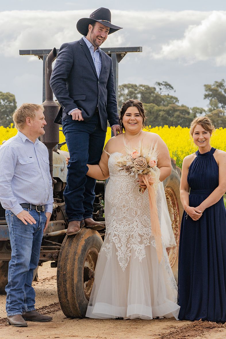 A wedding party poses together near a tractor with vibrant yellow canola fields in the background.