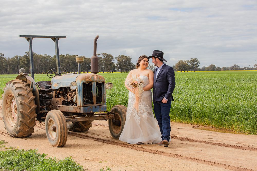A rustic country wedding shoot featuring a tractor and couple in formalwear on a dirt road surrounded by green fields.