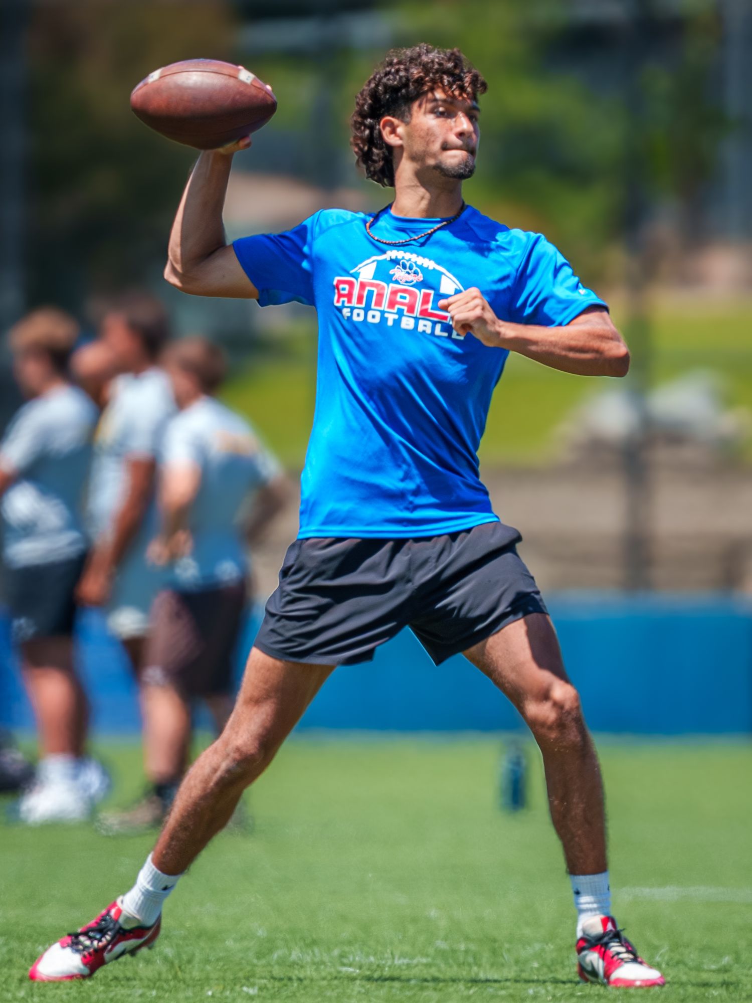 Football player in a bright blue shirt throws a pass during a summer practice session on an outdoor field.