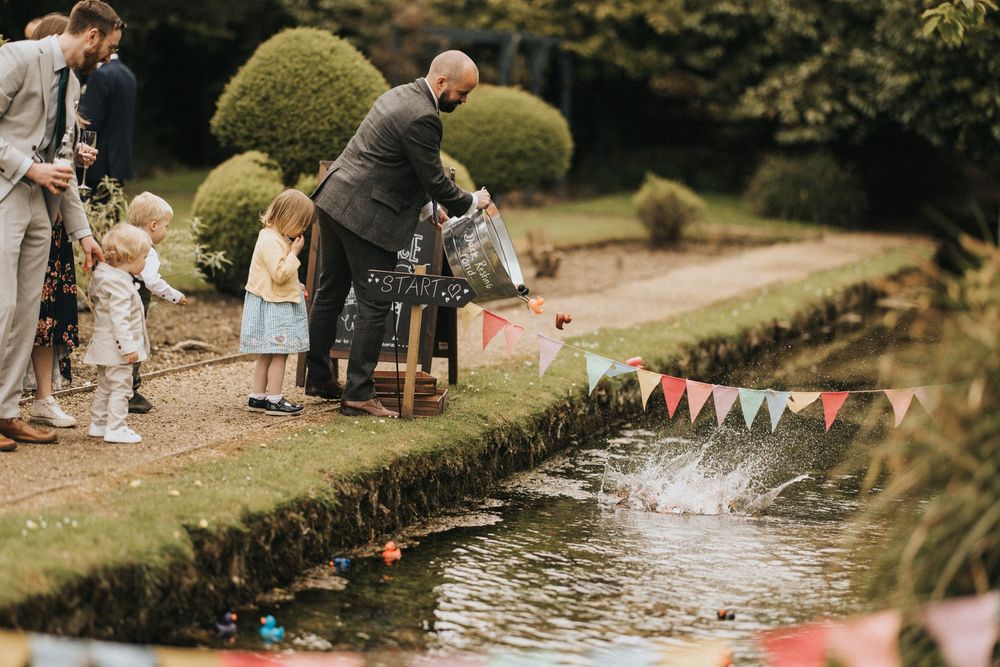 Standing around the edge of a pond at an outdoor party with festive bunting decorations, people gather to release paper boats.