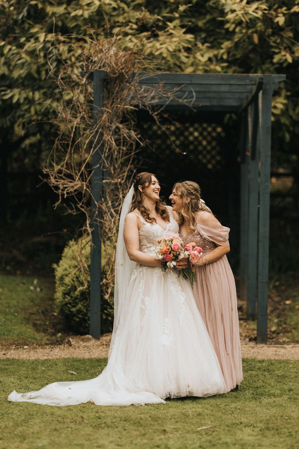 Two women in evening gowns share a joyful moment under a garden arbor decorated with dried flowers at an outdoor celebration.