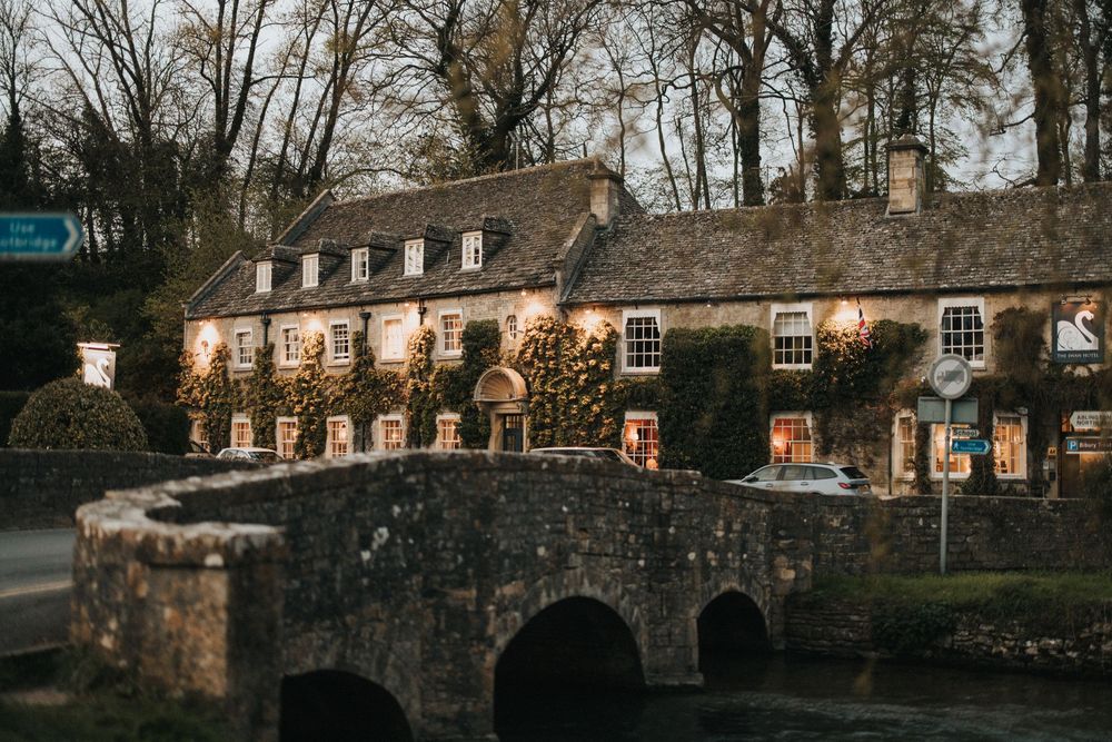 Historic Cotswold stone cottages with warm glowing windows beside an old stone bridge at dusk in Bibury, England.