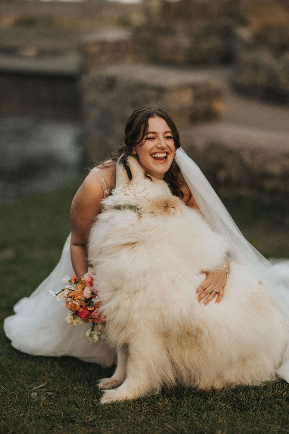 A bride in a flowing white gown laughs while laying in the grass during an outdoor wedding photoshoot.